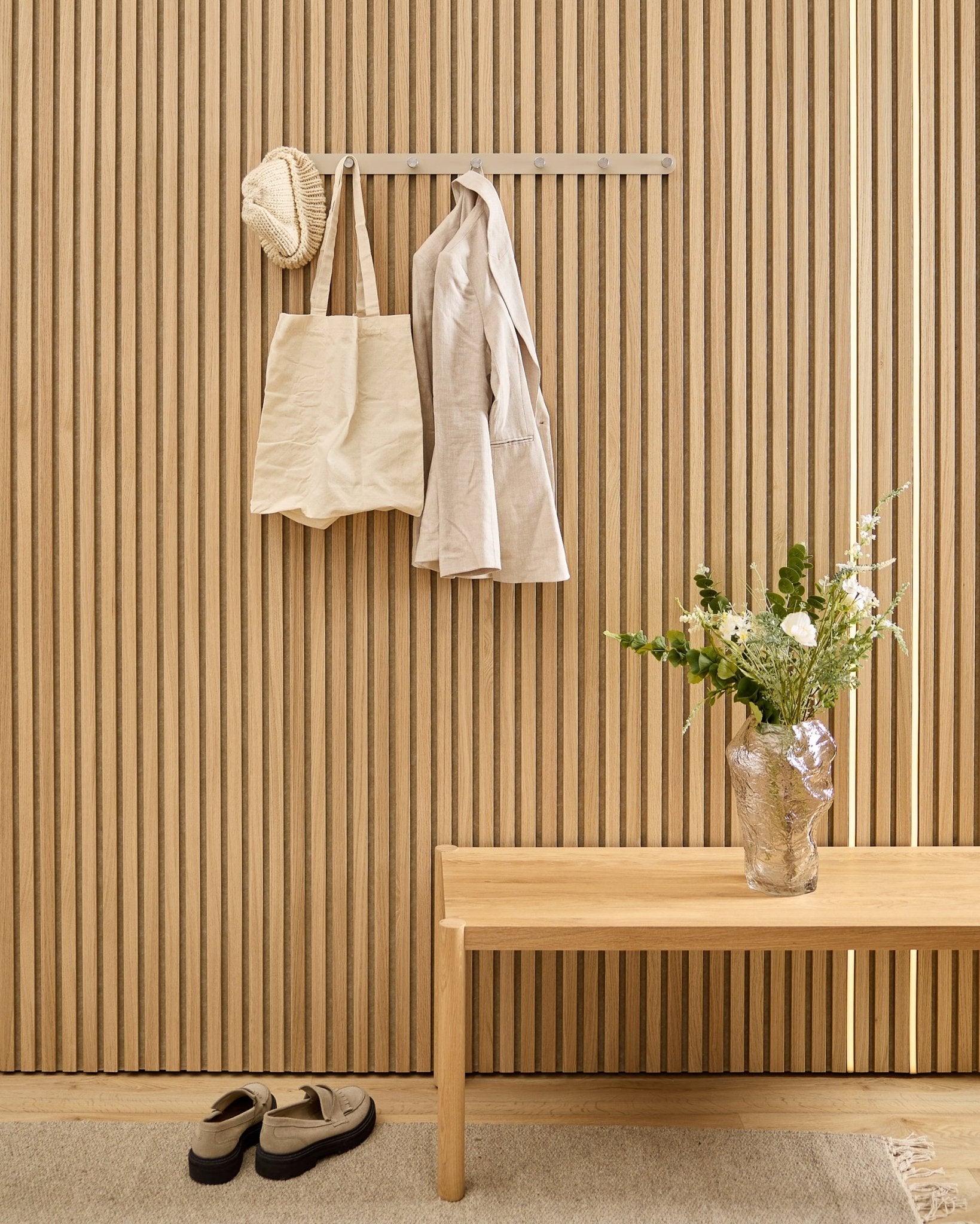 Un bolso beige, un sombrero y una camisa cuelgan del perchero de pared WoodUpp Norr sobre unos zapatos, con un banco y un jarrón junto al panel acústico de lamas a rayas.